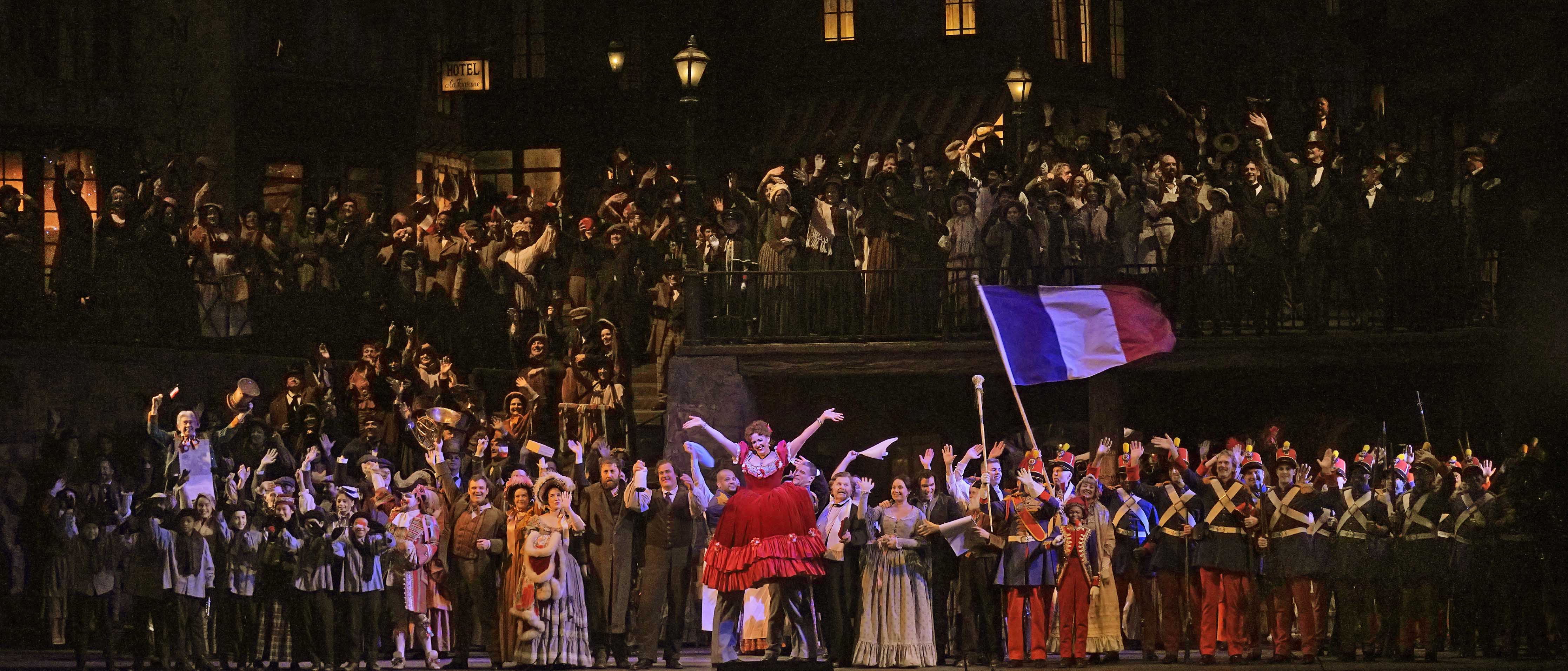 From a Met Opera production of "La Boheme," a large cast cover the stage, smiling and holding a French flag. Two men carry a woman in red at the center of the stage.