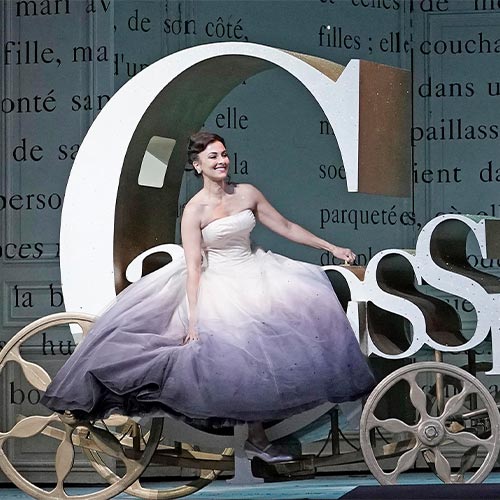 From the Met Opera's 2021 production of "Cinderella," Cinderella in a white ballgown riding a chariot pulled by four actors dressed as white horses. The chariot spells "carrosse," and the walls and horses'  suits have the words from the French book on them.